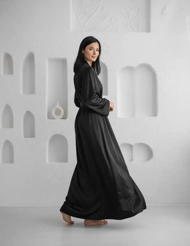 Woman in a long black dress standing against a white wall with geometric shapes. Zaine luxury black floor-length satin robe for women with a sophisticated wrap design, long sleeves and elegant premium satin finish.