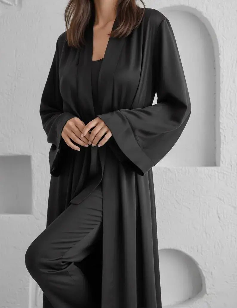 Person wearing a black long-sleeve dress against a white background. Zaine luxury black floor-length satin robe for women with a sophisticated wrap design, long sleeves and elegant premium satin finish.