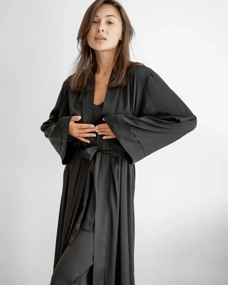 Woman wearing a black robe on a white background. Zaine luxury black floor-length satin robe for women with a sophisticated wrap design, long sleeves and elegant premium satin finish.