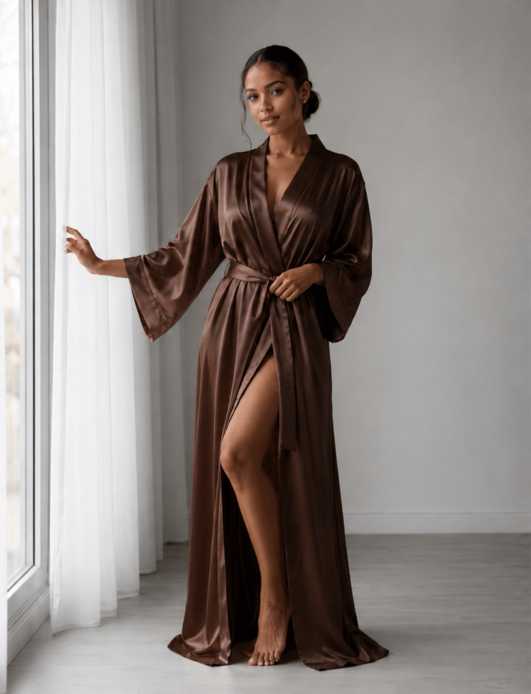 Woman wearing a brown satin robe standing by a window. Zaine Espresso Elegance luxury floor-length satin robe for women featuring beautiful elasticated sleeves, a flattering wrap silhouette and silky premium satin fabric.
