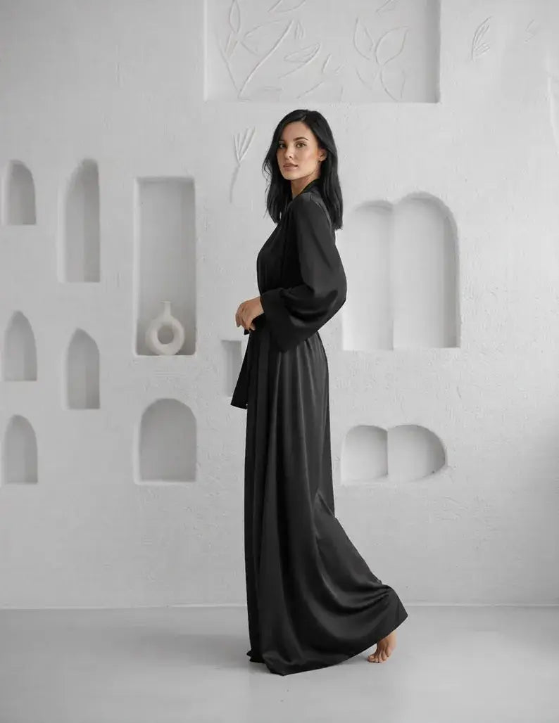 Woman wearing a black dress standing against a white wall with geometric shapes. Zaine luxury black floor-length satin robe for women with a sophisticated wrap design, long sleeves and elegant premium satin finish.